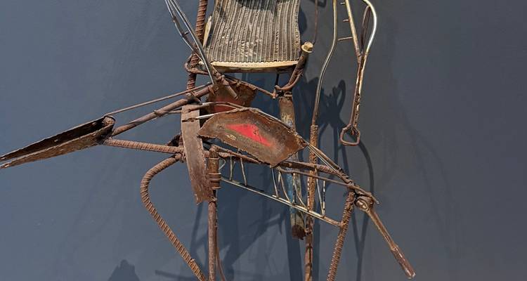 Primer plano de una escultura de metal contemporánea hecha con piezas de chatarra soldada, exhibida contra una pared oscura de galería.