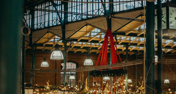 Interior de un mercado histórico decorado con luces colgantes y adornos festivos bajo un techo de hierro y cristal.