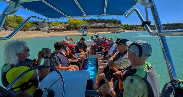 Tour group wearing life jackets seated in an open motorboat on turquoise water near a sandy shore and beach huts.