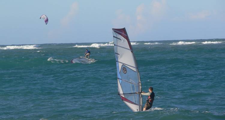 Windsurfer in action on choppy teal waves with a kite-surfer in the distance under a clear sky.