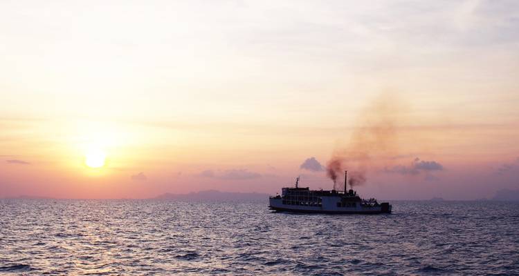 Silhouet van een veerboot die rook uitstoot over een kalme zee bij zonsondergang.