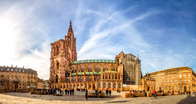 Large panorama de la Cathédrale de Strasbourg dominant sa place centrale animée sous un ciel bleu.