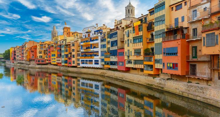 Las coloridas fachadas ribereñas de Girona reflejándose en aguas tranquilas bajo un cielo azul radiante.