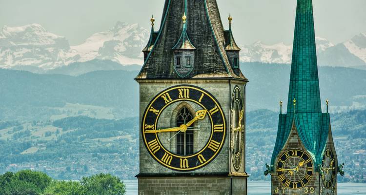 Großer mittelalterlicher Glockenturm mit alpinem Seepanorama im Hintergrund und grünem Spitzendach in der Nähe.