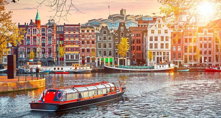 Kanaalscène in Amsterdam bij zonsondergang met een rondvaartboot met glazen dak dat langs kleurrijke huizen met trapgevel glijdt.