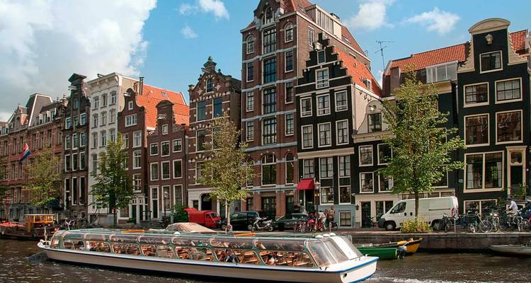 Een rondvaartboot en smalle grachthuizen liggen langs een Amsterdams waterkanaal onder een wolkige hemel.