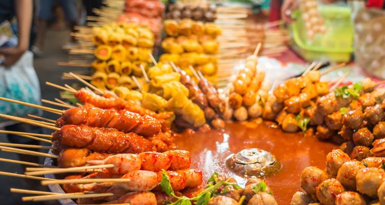 Assortiment coloré de brochettes grillées disposées autour d'une sauce épicée dans un marché de nuit thaïlandais animé.