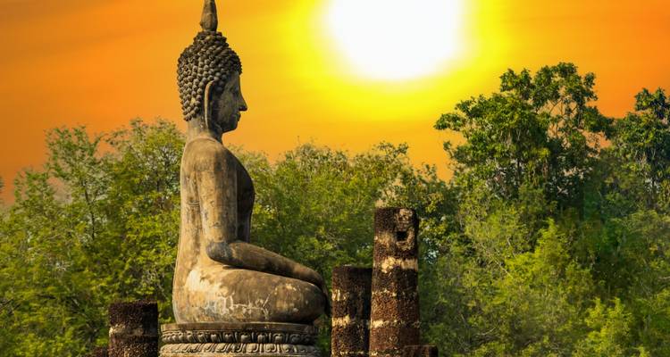 Statue de Bouddha en pierre ancienne de profil sur un coucher de soleil orange éclatant dans un parc verdoyant.
