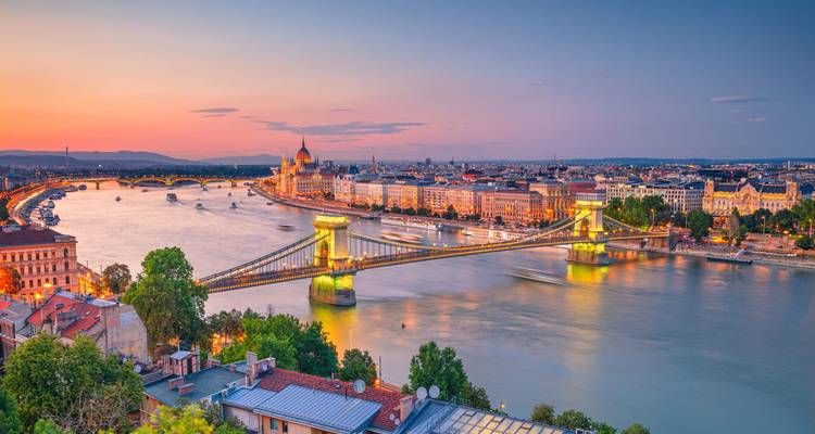 Panoramic sunset view over Budapest featuring the Chain Bridge spanning the Danube and the illuminated Parliament building.
