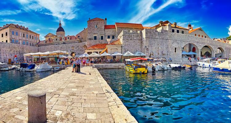 Stone harbour walls of a historic city with cafés and boats reflecting on vivid blue water.