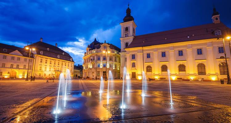 Illuminated baroque square with fountains spraying water against a twilight sky.