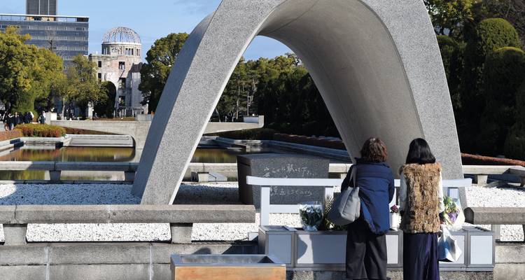 Visitors paying respects at Hiroshima Peace Memorial Cenotaph with Atomic Bomb Dome visible.