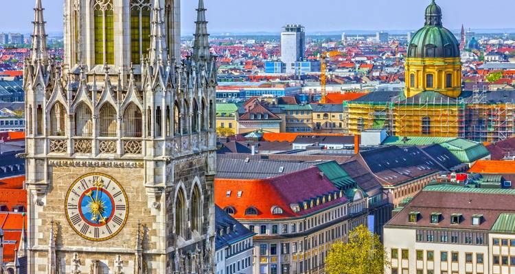 Tagsüber aufgenommene Stadtansicht von München mit dem neugotischen Turm des Neuen Rathauses, das über farbenfrohe Dächer hinwegsieht.