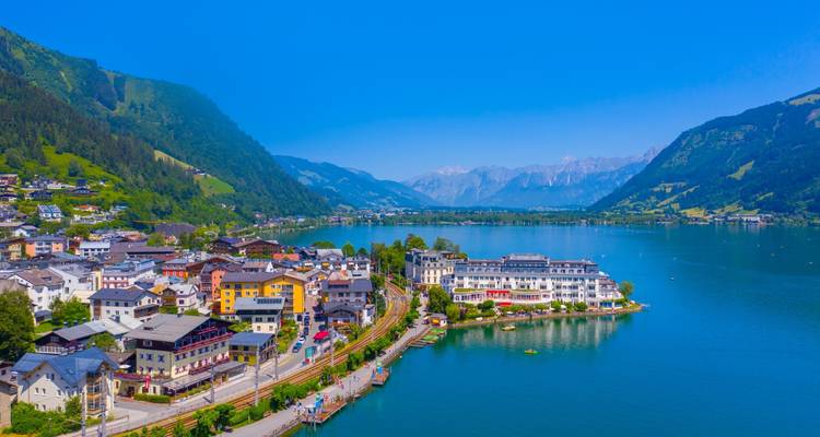 Luftaufnahme des Ortes Zell am See im Sommer, der sich an einen türkisfarbenen Alpensee schmiegt, mit hohen Bergen im Hintergrund.