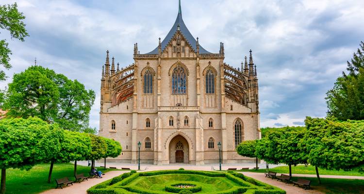 Vooraanzicht van St. Barbara's Church in Kutná Hora omgeven door verzorgde tuinen en weelderig groen onder een bewolkte lucht.