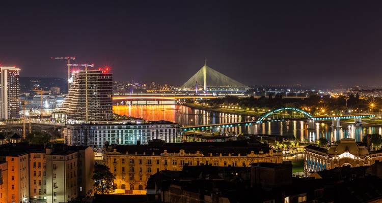 Nächtliches Panorama einer Flussstadt mit einer beleuchteten Schrägseilbrücke und Spiegelungen im Wasser.