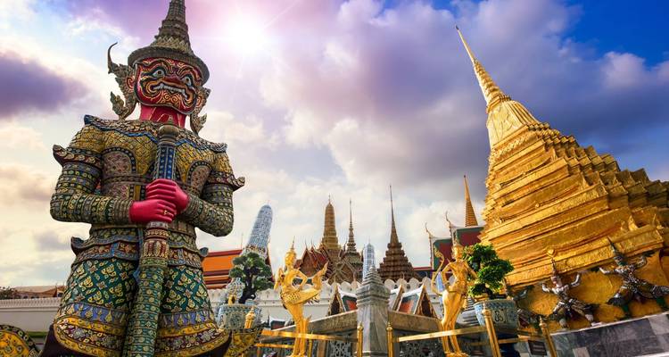 Complejo del Gran Palacio en Bangkok con una gigantesca estatua guardiana y una estupa dorada bajo un cielo vibrante.