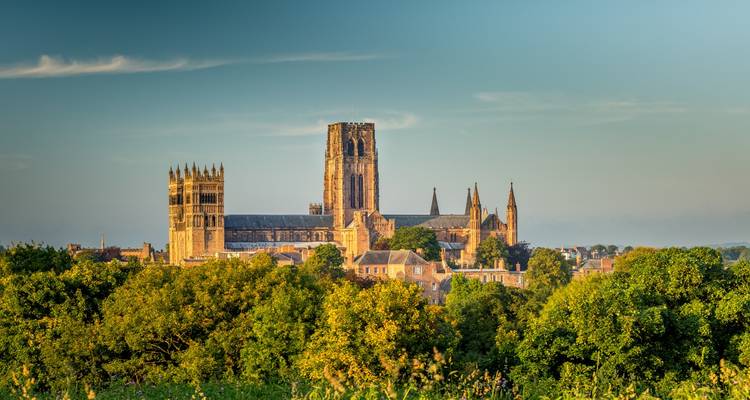 De goudgeel verlichte Durham Cathedral en het kasteel die boven de boomtoppen en historische daken uitsteken tegen een heldere avondhemel.