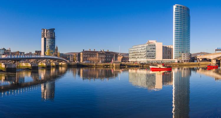 Moderne skyline van Belfast met bruggen, rode boten en een kalme rivier die weerspiegelt onder een helderblauwe hemel.
