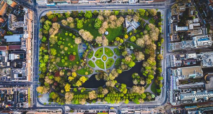 Drone-opname van bovenaf van een rechthoekig stadspark met vijvers, wandelpaden en kleurrijke bomen omringd door drukke straten.