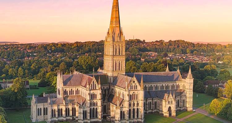 Aerial sunrise view of Salisbury Cathedral amid lush English countryside with warm golden light.