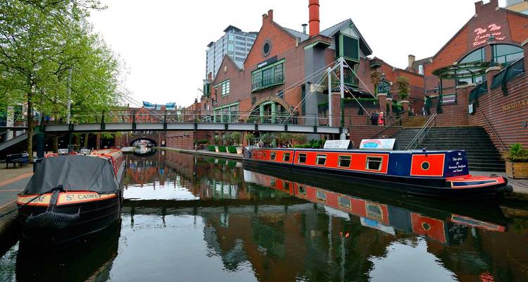 Colorful narrowboats sit on a calm urban canal lined by red-brick warehouses and a footbridge in the city center.