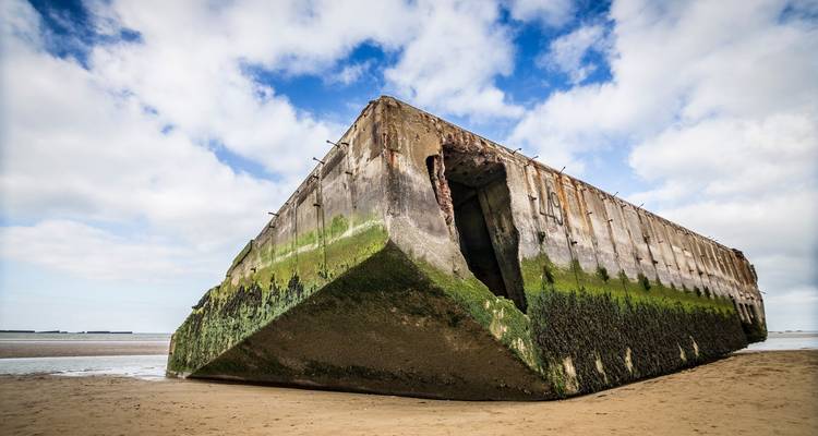 Enorme caisson del puerto artificial Mulberry varado en una playa de Normandía con franjas verdes cubiertas de musgo