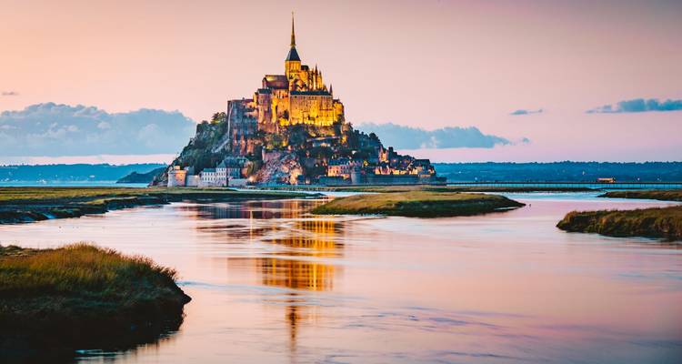 Mont Saint-Michel iluminado por el oro reflejado en canales de marea al atardecer con un cielo pastel