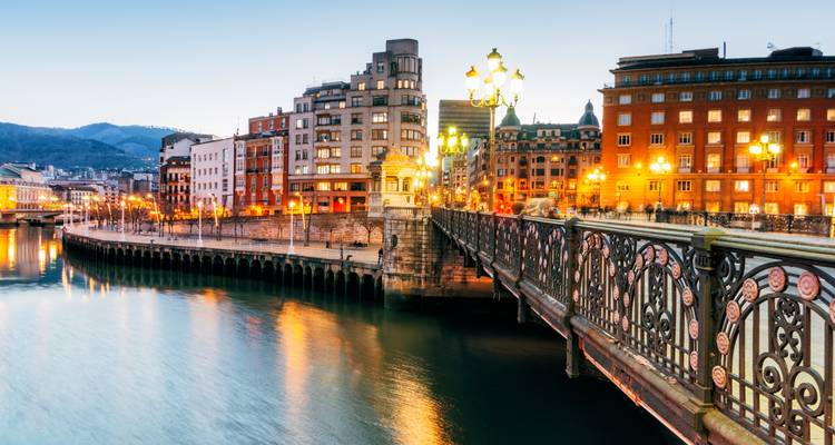 Evening view of ornate bridge and riverside buildings glowing under streetlights in Bilbao.