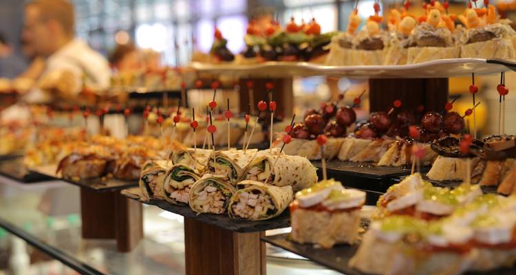 Assorted pintxos skewered on small pieces of bread arranged on tiered platters inside a lively tapas bar.