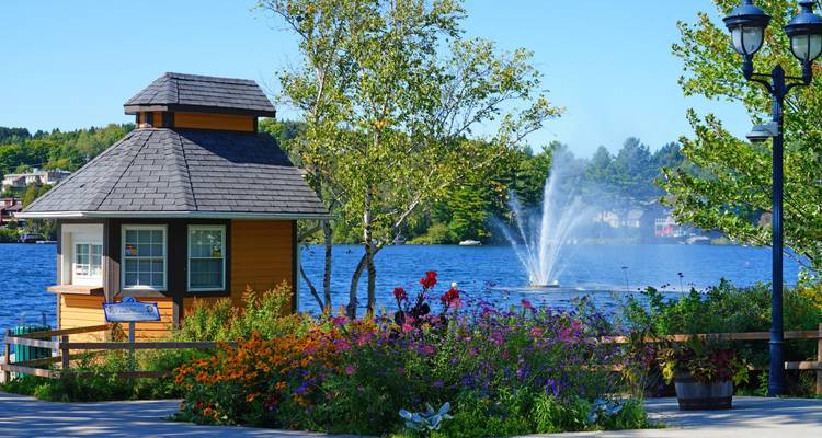Colourful flower garden and lake fountain beside a small wooden pavilion on a sunny summer day.