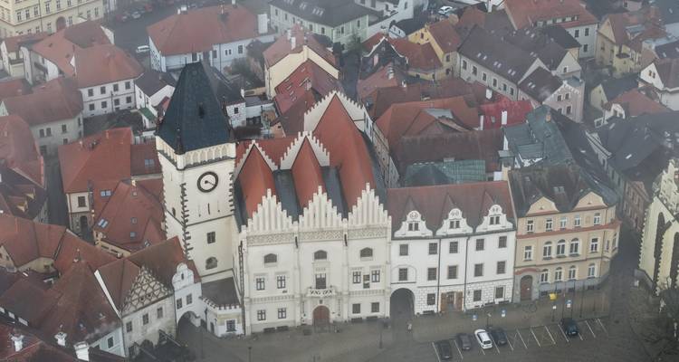 Vista aérea nebulosa de edificios históricos con techos rojos agrupados alrededor de una torre del reloj blanca en la plaza de un pueblo checo.