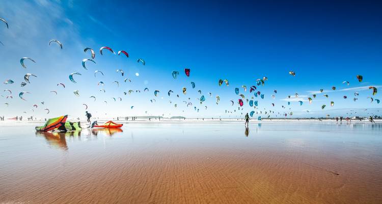 Breiter Sandstrand mit Dutzenden bunten Drachen, die an einem klaren, blauen Tag in der Luft aufsteigen, während Kitesurfer ihre Ausruestung vorbereiten.