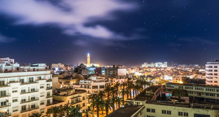 Nachtpanorama einer modernen Stadt mit dem beleuchteten Minarett der Hassan-II-Moschee, das sich über palmenbepflanzte Boulevards unter einem sternenklaren Himmel erhebt.