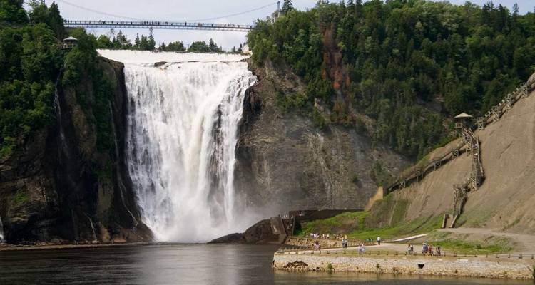 Powerful Montmorency Falls plunging beside a forested cliff with visitors on the shoreline and suspension bridge above.
