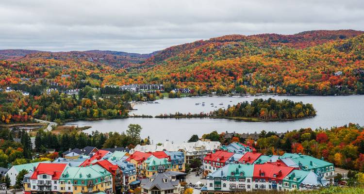 Aerial autumn view of Mont-Tremblant village with colorful roofs beside a lake surrounded by forested hills ablaze in fall colors.