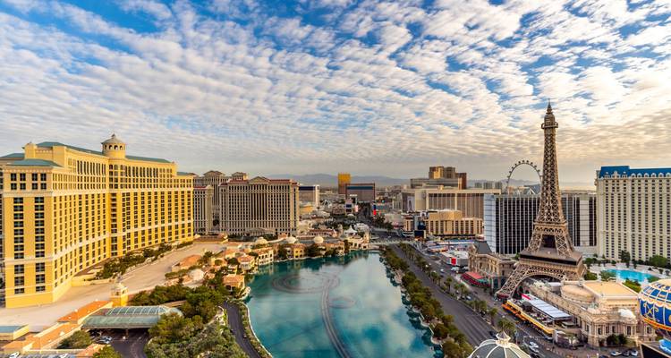 Expansive daylight view of the Las Vegas Strip with Bellagio fountains, Eiffel Tower replica and High Roller under patterned sky.