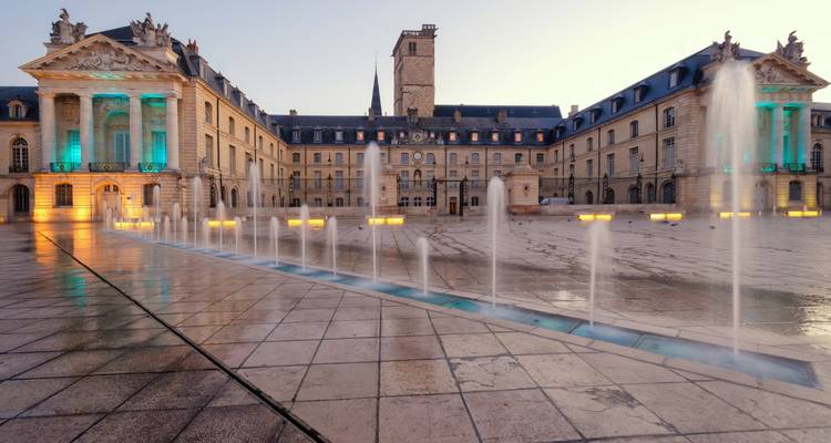 Grand Place Royale à Dijon avec des fontaines projetant des jets d'eau devant le majestueux Palais des Ducs illuminé au crépuscule.