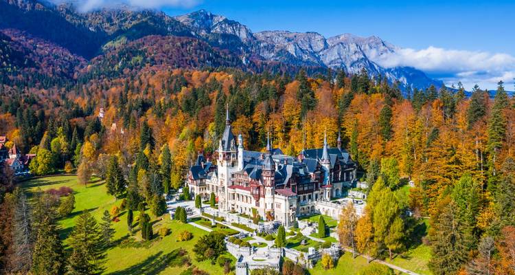 Aerial autumn view of Peles Castle nestled in colourful Carpathian forest with mountain backdrop.