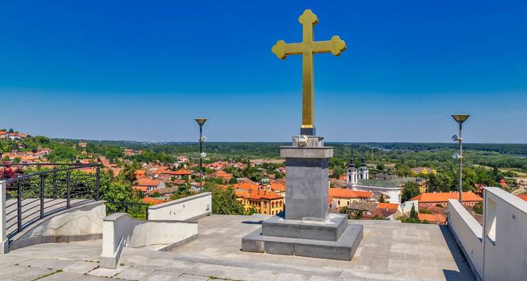 Large golden cross monument overlooking rooftops and green plains of Sremski Karlovci.