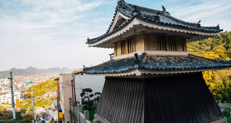 Traditional Japanese turreted structure on a hillside overlooking a distant coastal city and green forested slopes.