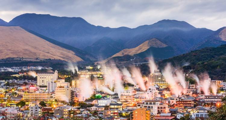 Columnas de vapor blanco se elevan desde numerosas casas de baños en el pueblo termal de Beppu contra fondos montañosos al atardecer.