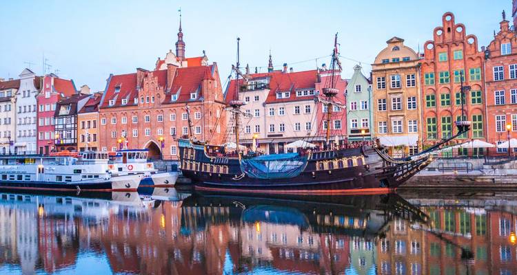 Edificios históricos de colores vivos en el paseo marítimo y un barco antiguo al estilo de galeón reflejados en el río Motława al amanecer.