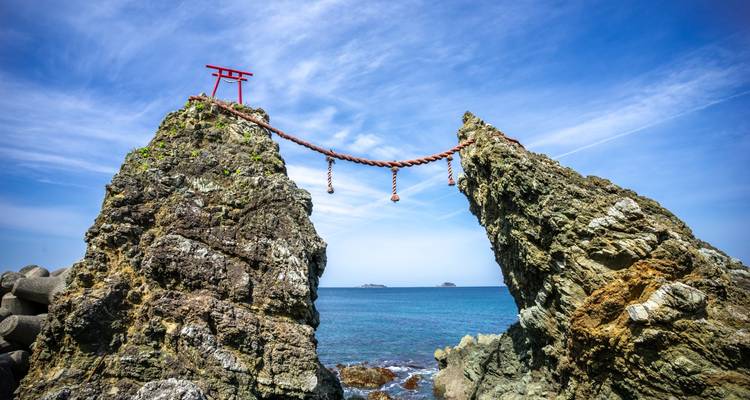 Twin sea-bound rocks connected by sacred rope beneath a bright blue sky and clear ocean.