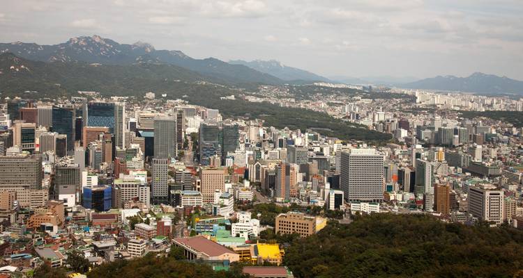 Expansive skyline of a modern Asian city backed by forested mountains under hazy light.