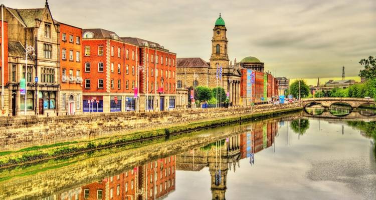Colorful historic buildings along river with reflections and bridge, under moody sky