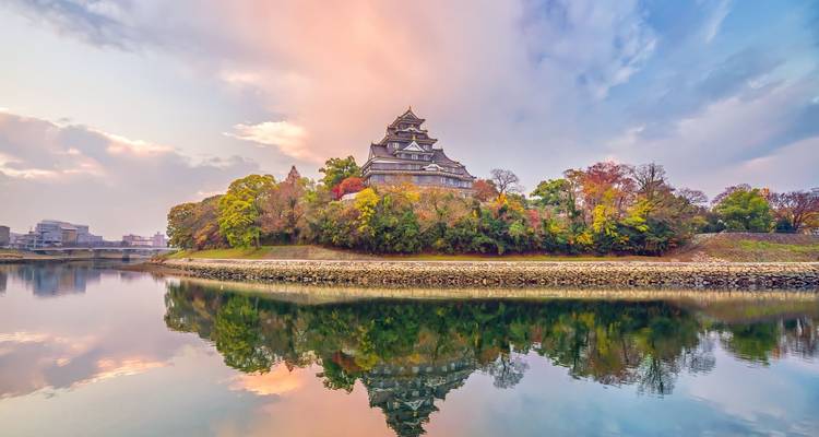 Traditional Japanese castle sitting on a tree-covered island reflected perfectly in a calm river at sunset.