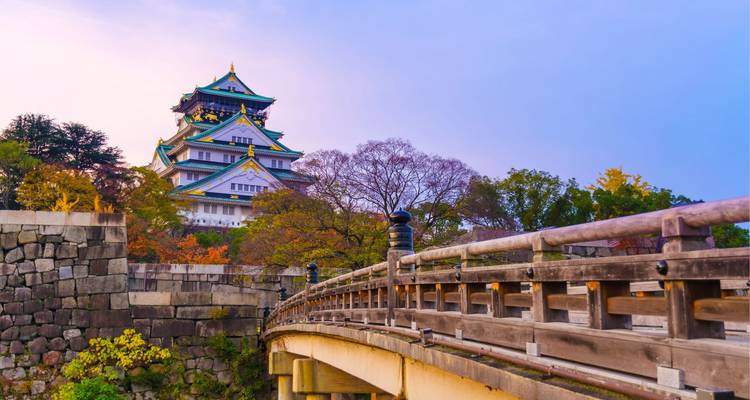 Iconic Osaka Castle rises behind an arched wooden bridge surrounded by colourful autumn foliage in soft evening light.