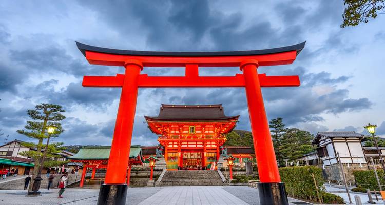 Bright vermilion torii gate standing before an illuminated shrine with dramatic evening clouds overhead.