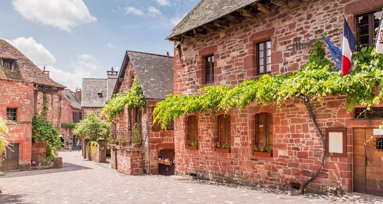 Les petites maisons pittoresques en grès rouge, drapées de vignes, bordent les ruelles pavées de Collonges-la-Rouge sous un ciel lumineux.